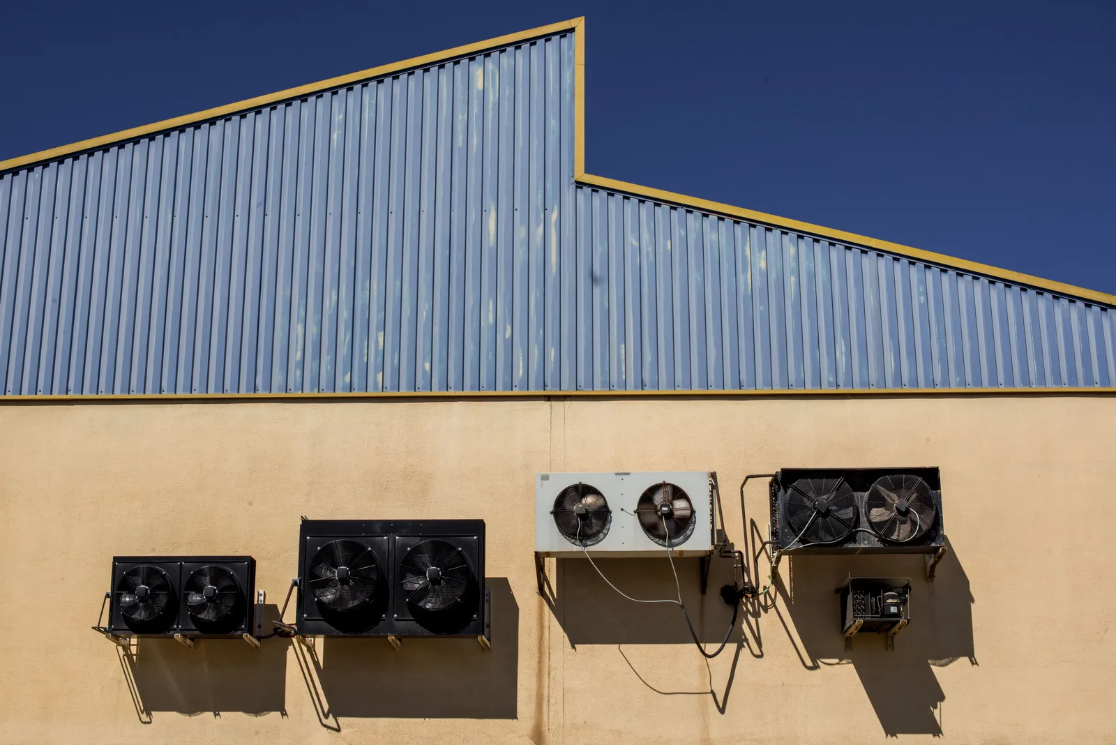 A row of industrial air conditioning condensing units mounted along a warehouse wall under a clear sky
