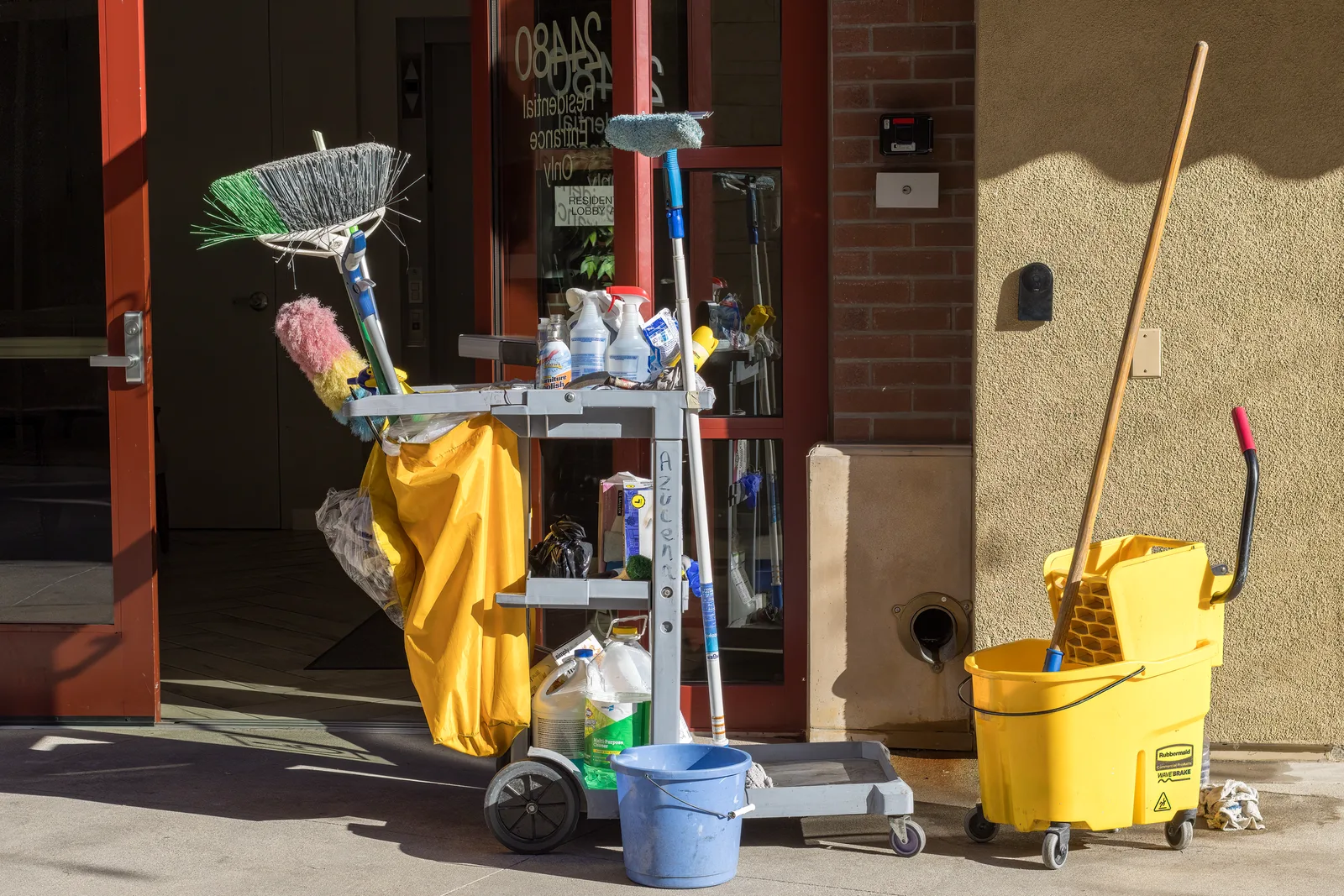 Buckets, brushes, mops and cleaning bottles staged on the ground outside a building entrance.