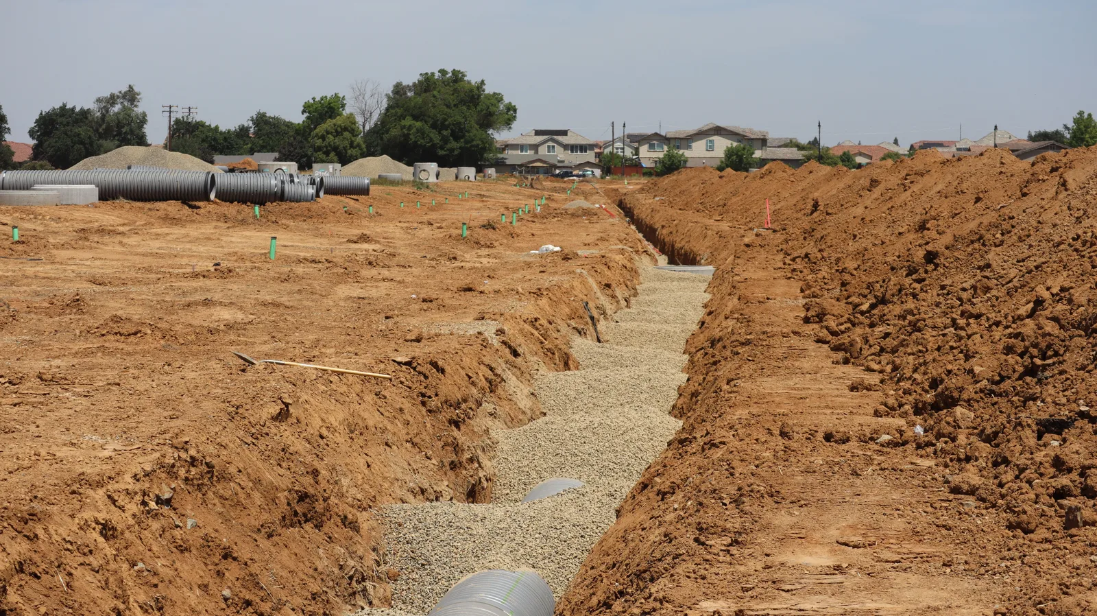 A wide view of a suburban construction site with a long open trench and pipe stacked alongside it