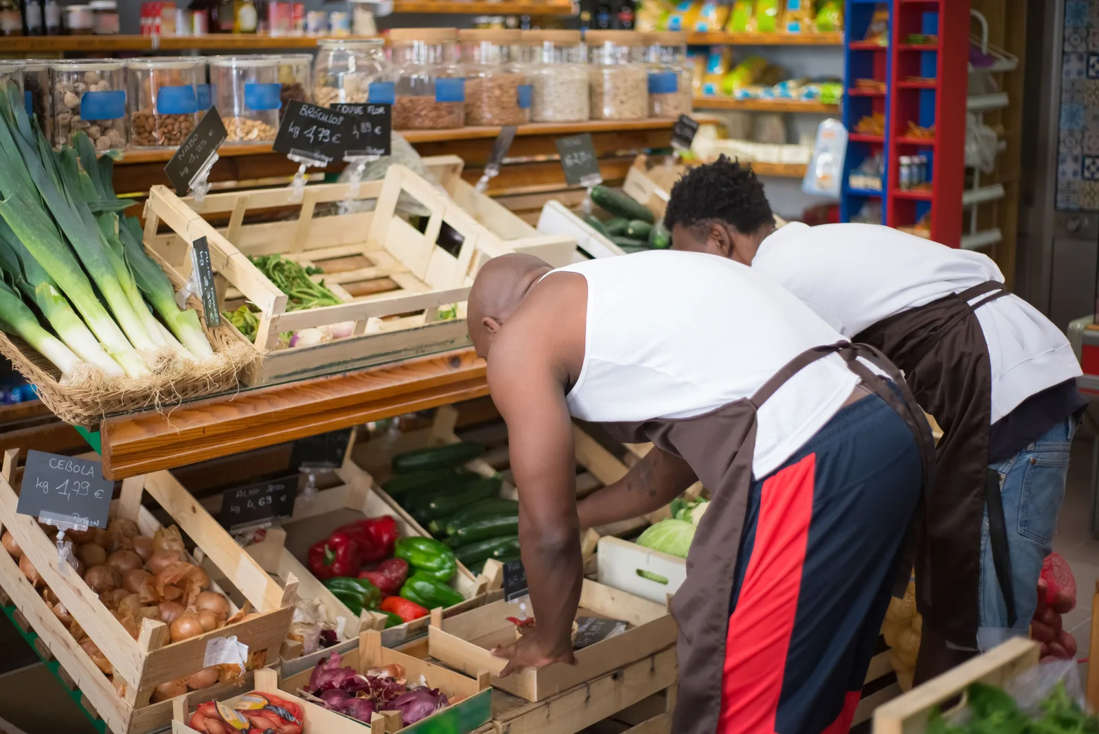 Two grocers in aprons arranging vegetables on a produce display inside a store.