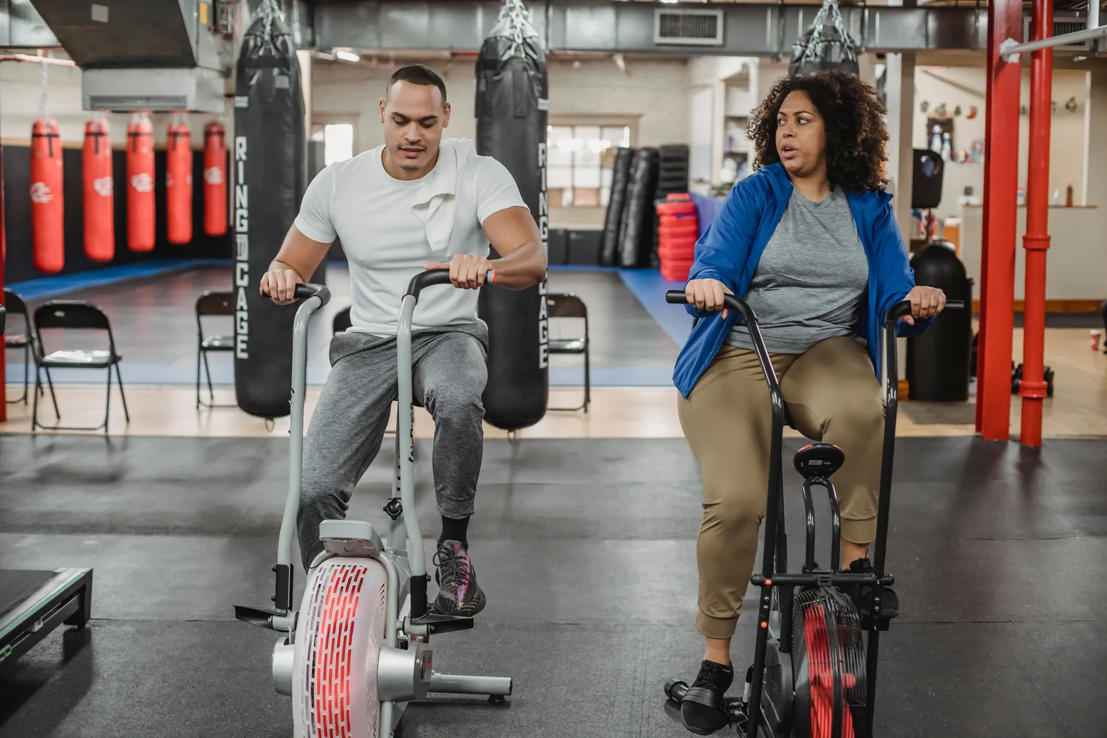 Two gym members training side by side on air bikes on a gym floor, with punch bags along the wall behind