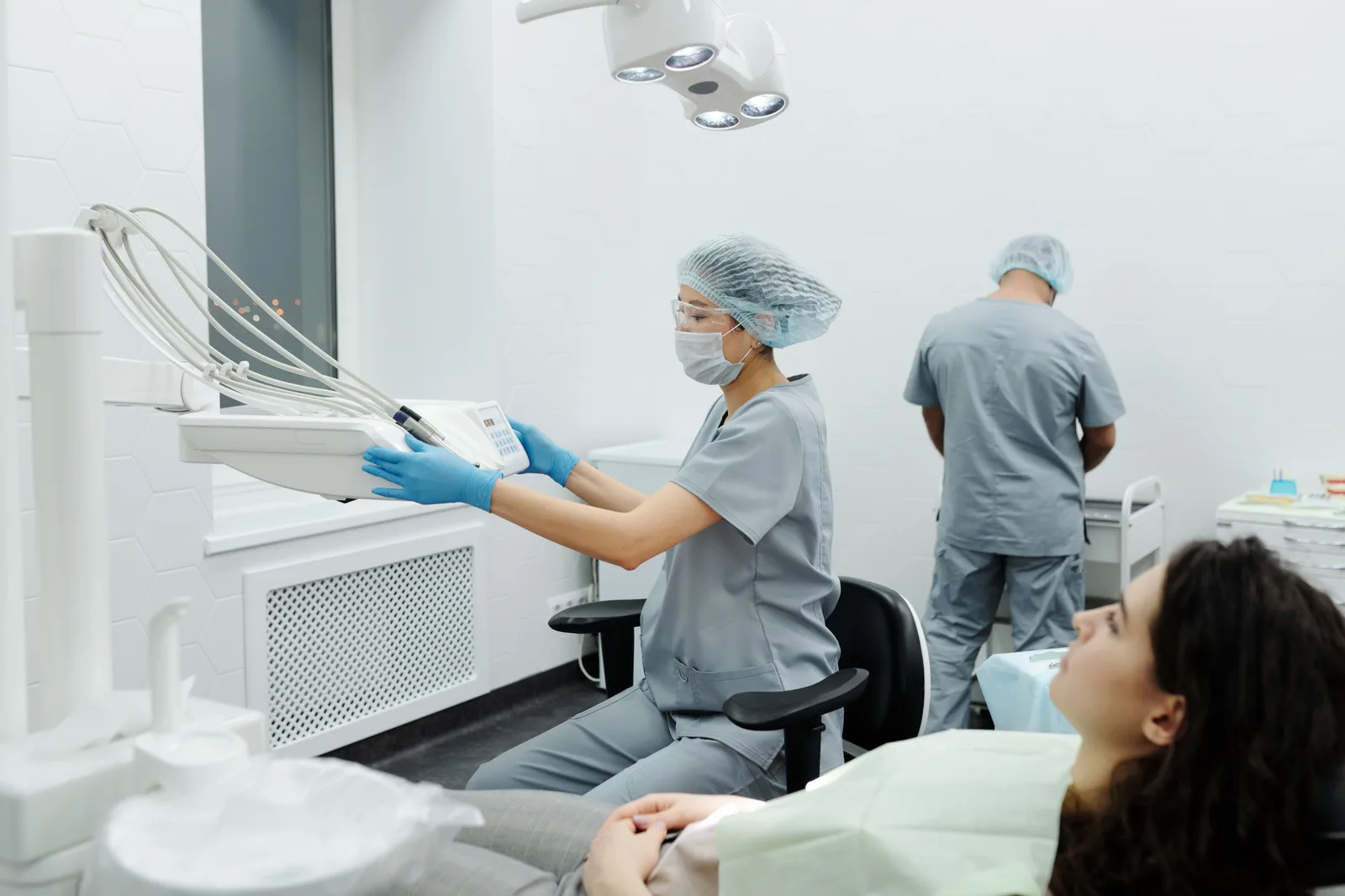 A dental assistant in scrubs and a surgical cap adjusting an instrument arm beside a patient reclined in a dental chair, with a second clinician working at the counter behind