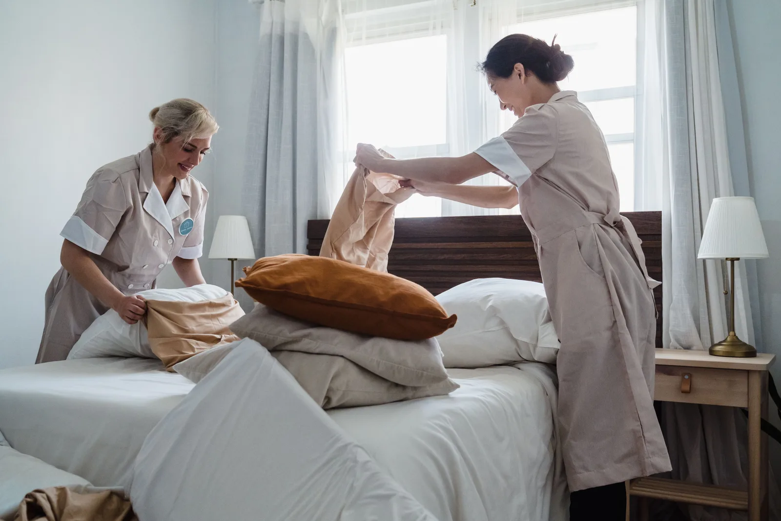 Two housekeepers in uniform making up a bed with fresh linen in a guest room.