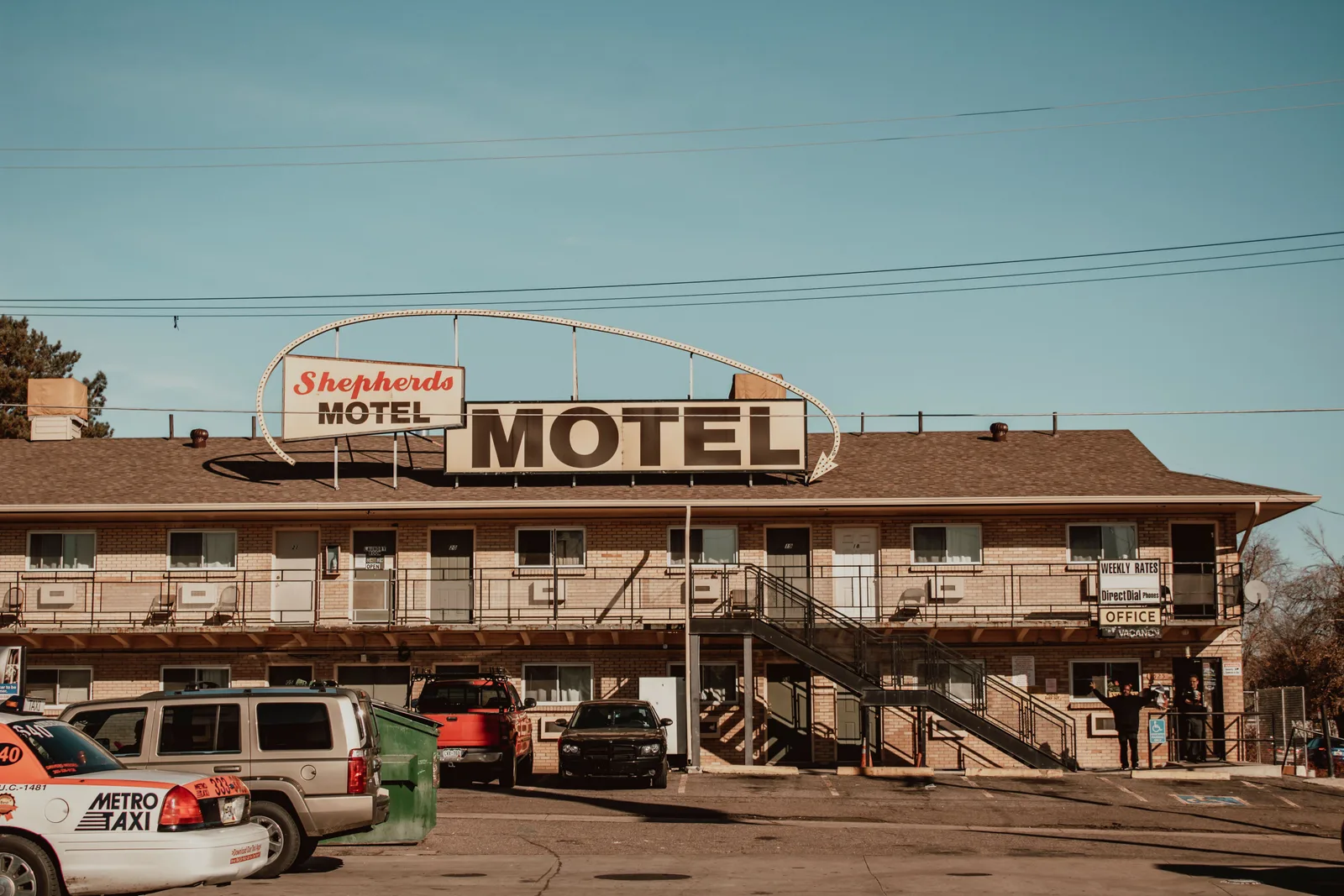 A two-story independent motel with a large rooftop sign, an office window advertising weekly rates and vacancies, and cars parked in front of the rooms.