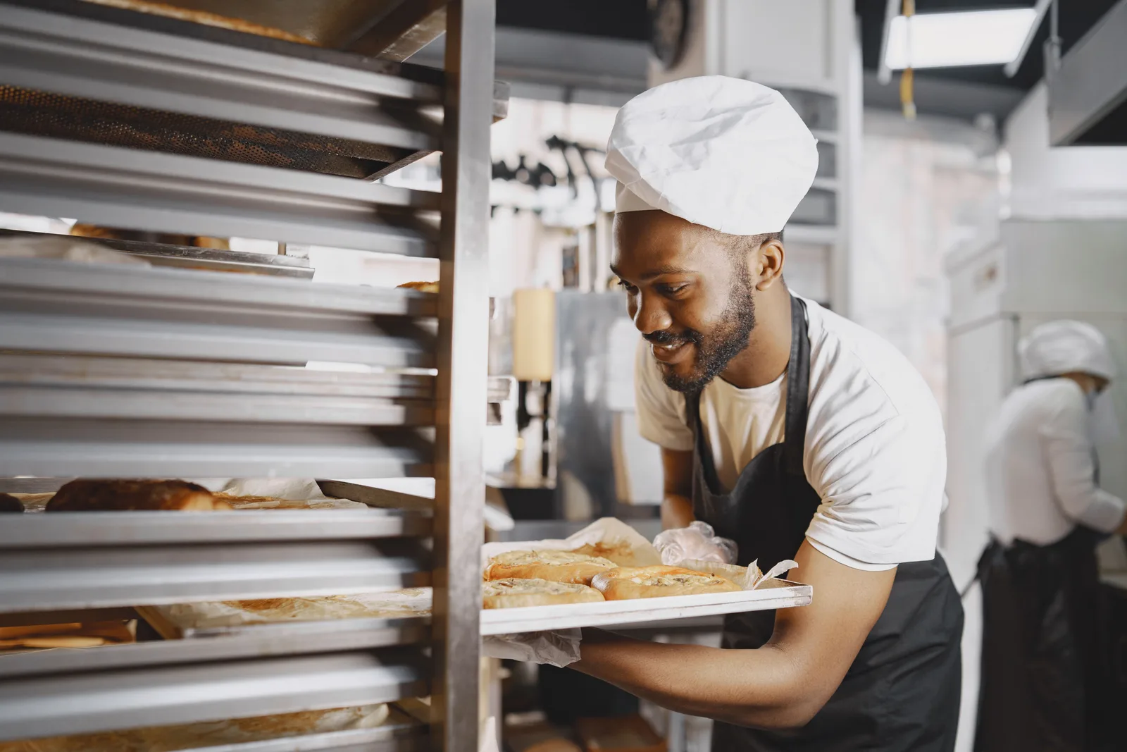 A baker carries a tray of freshly baked bread past a rack in a commercial bakery.
