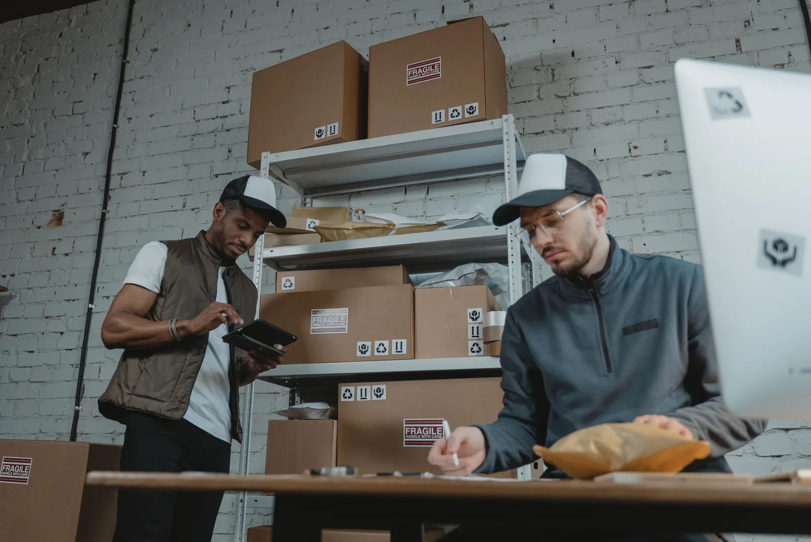 Two workers pack and label parcels on shelving in a small order fulfilment room.