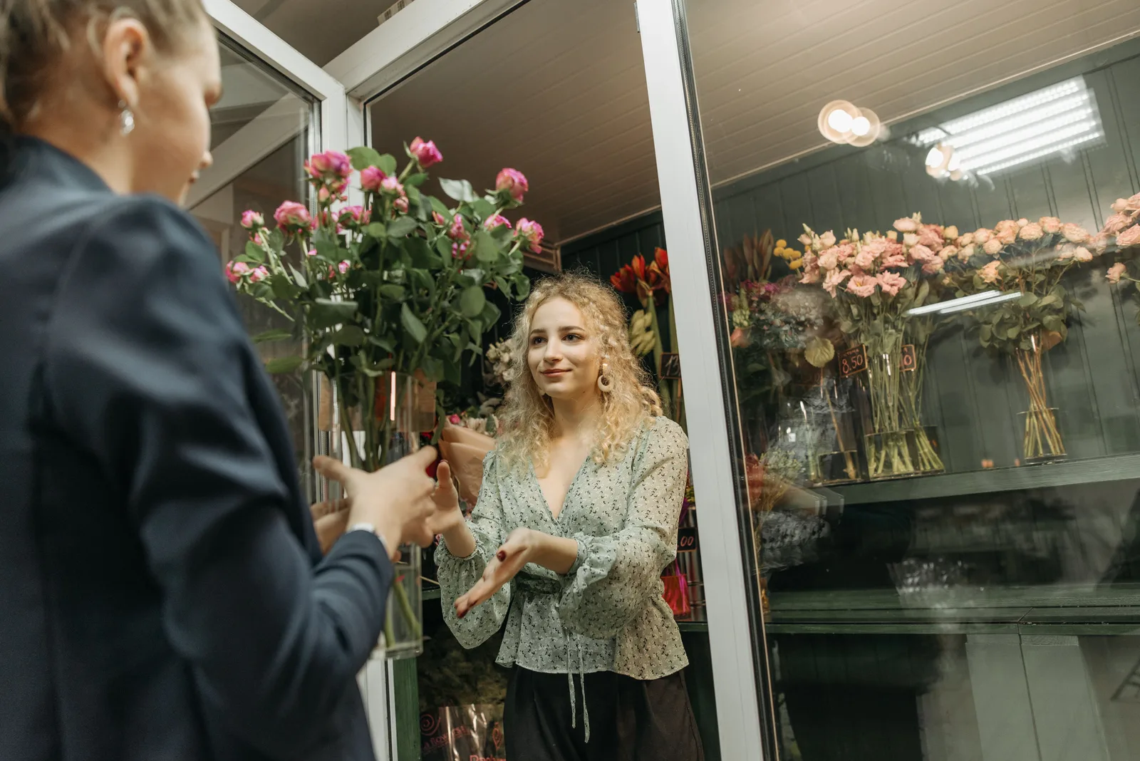 A florist passing wrapped stems across to a customer in her shop, with chilled display cabinets of flowers behind her.