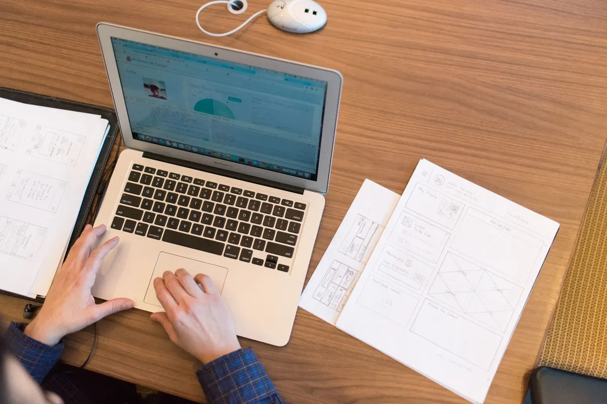 Overhead view of a person using a laptop trackpad with printed pages spread on a wooden desk beside it.
