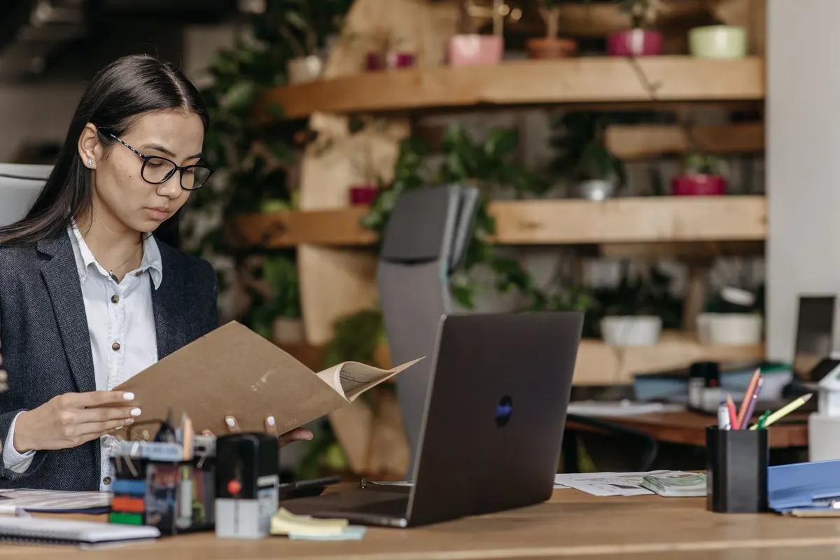 Woman in glasses and a gray blazer looking through a brown folder at a desk with a laptop, pen holder and sticky notes.