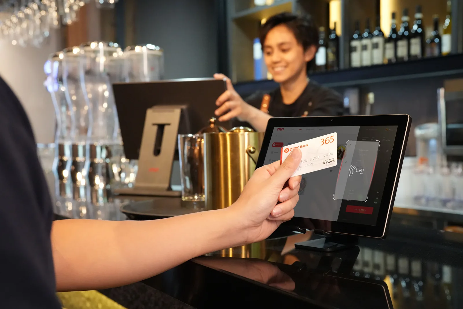 Customer tapping a bank card on a countertop payment terminal while a member of staff reaches across the bar