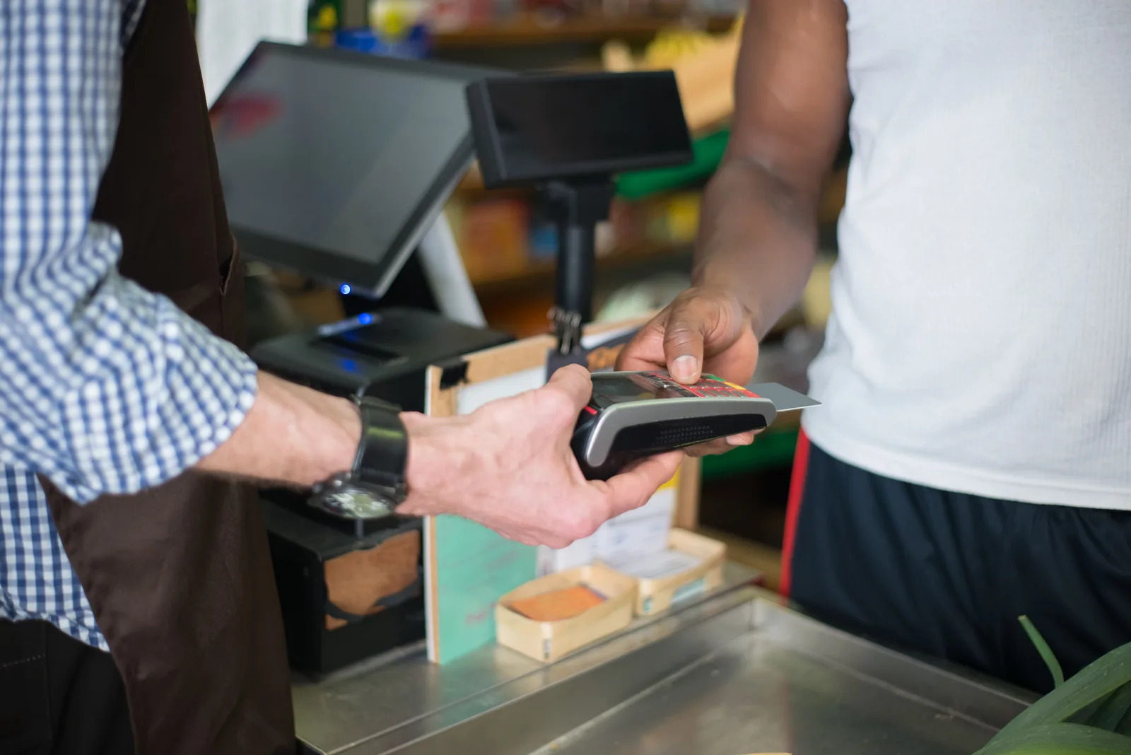 Hands passing a card across a shop counter to a portable terminal held by a clerk, beside a point of sale screen and receipt printer