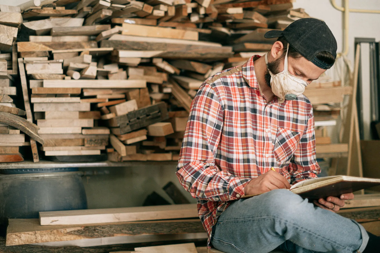 A man in a plaid shirt, cap and dust mask sitting in a woodshop writing on a board, with stacked offcuts and timber behind him