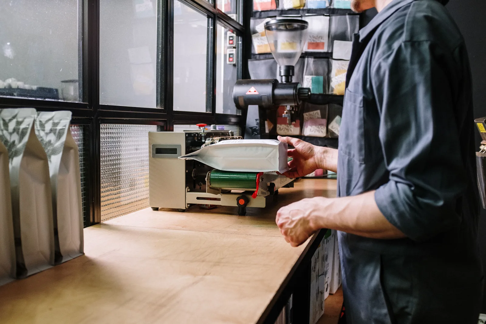 A worker in a small roastery sealing a filled coffee bag on a bench top packing machine.