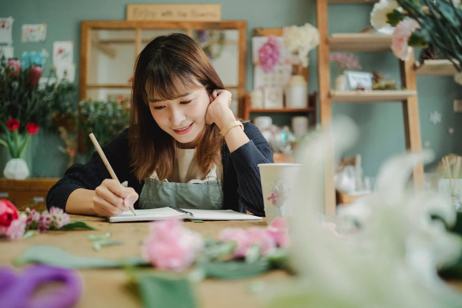 A florist in an apron writing in a notebook at the shop workbench surrounded by cut flowers and ribbon