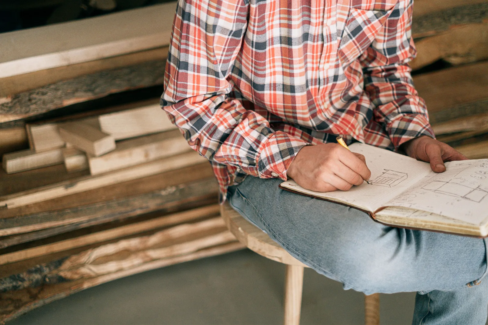 A joiner in a plaid shirt sketching a design in a notebook resting on their knee beside stacked timber