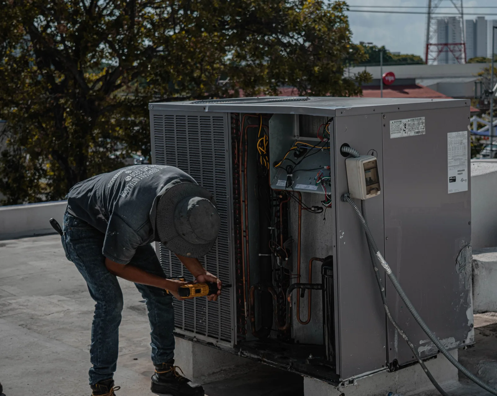 Technician leaning into an opened rooftop HVAC unit with a cordless drill, wiring and coils exposed