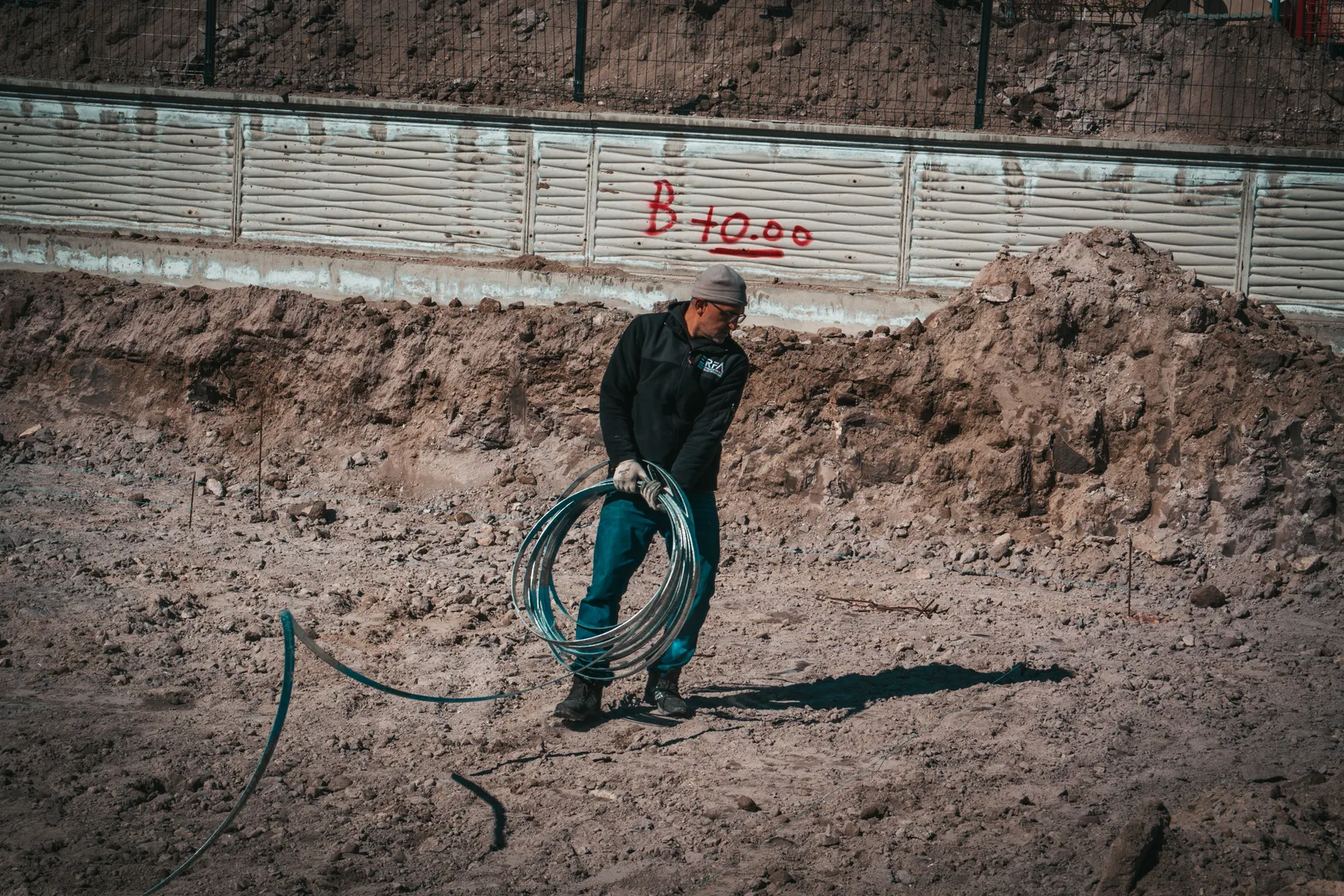 A construction worker carrying a heavy coil of electrical cable across an excavated site