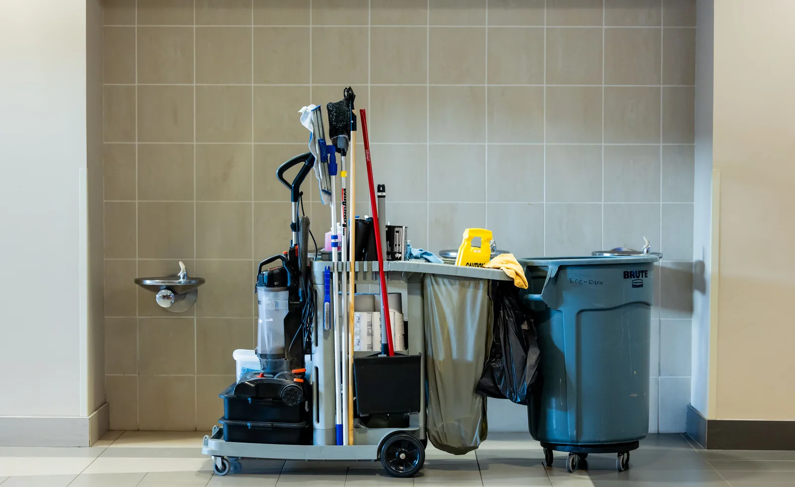 A professional janitorial cart loaded with mops, cloths, spray bottles and paper supplies in a building corridor.