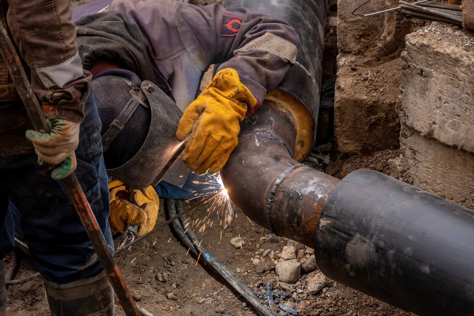 A worker in gloves welding a joint on a large steel pipe down in an excavation, with sparks flying