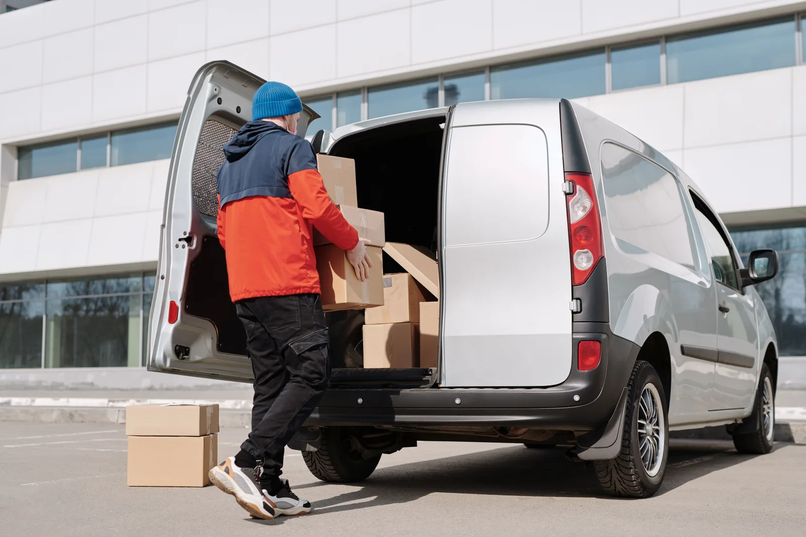 A man in a work jacket lifting cardboard boxes into the back of a small delivery van outside a commercial building