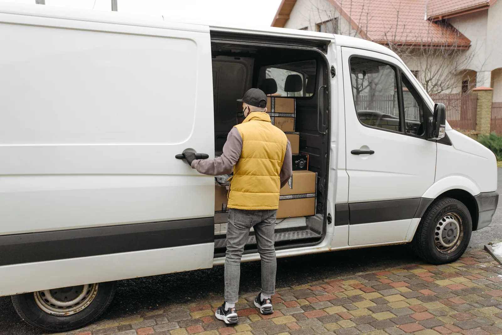 Driver loading parcels into the side door of a white delivery van parked outside a house