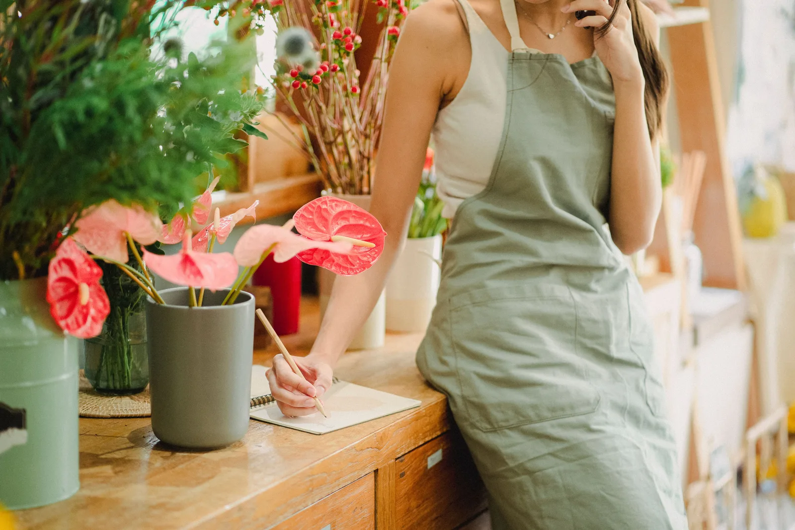 Florist writing in a notebook at her shop bench while holding a phone to her ear