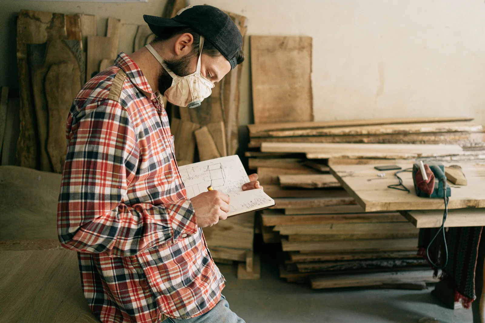 Craftsman in a dust mask reading a hand drawn plan in a workshop stacked with timber