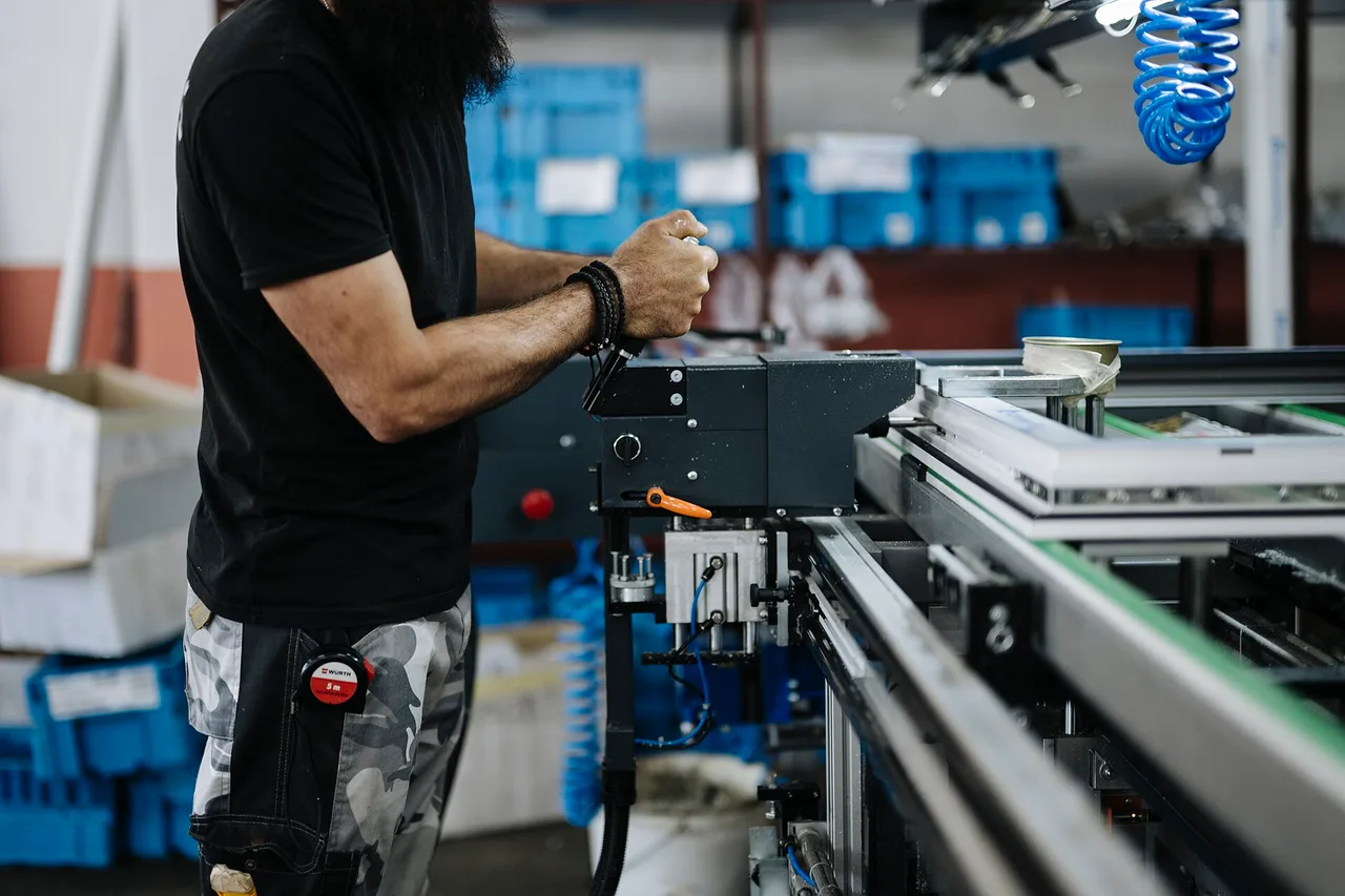 A worker operates a bench-mounted pneumatic press beside an aluminium conveyor, with stacked blue parts bins behind