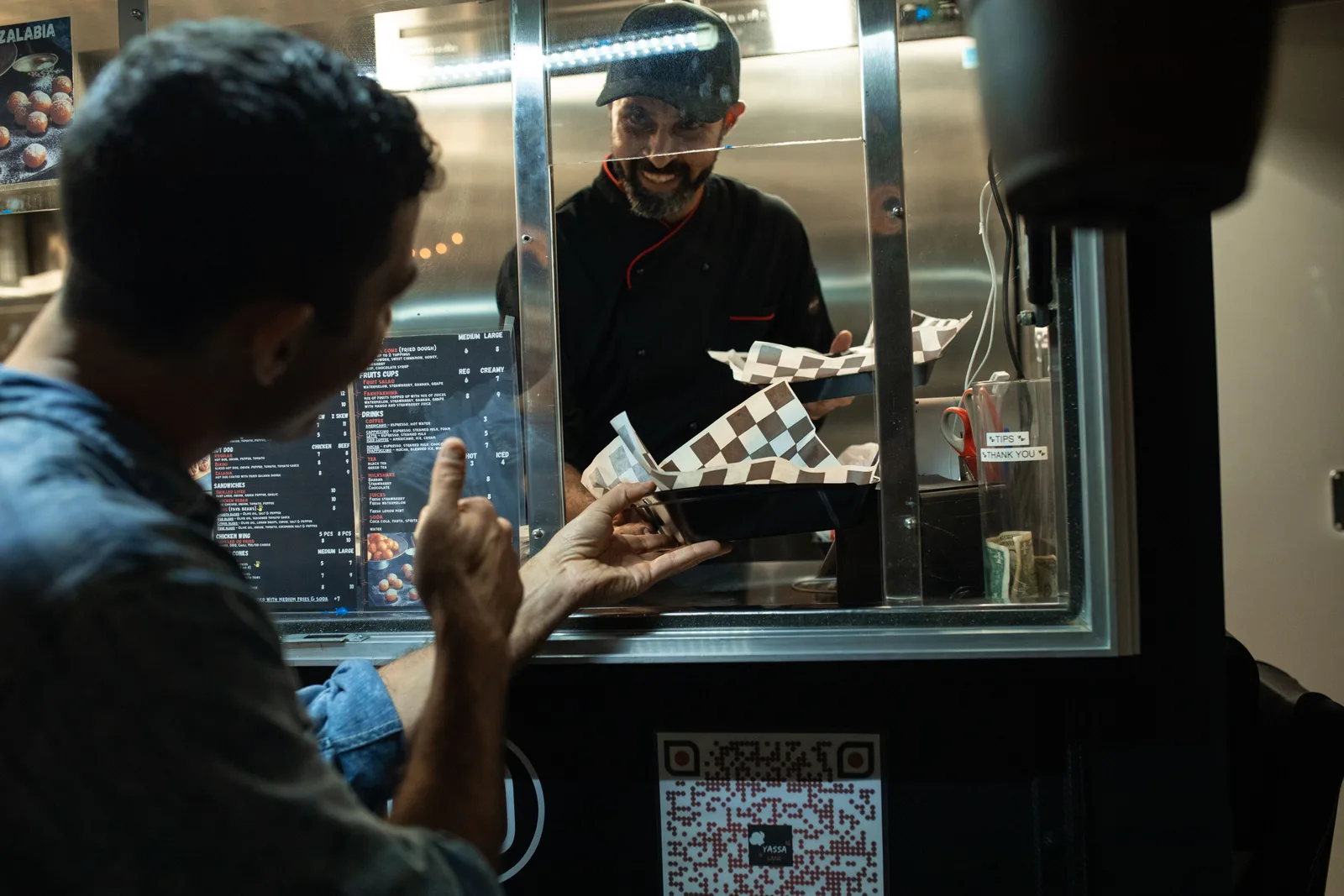 Customer taking a paper tray of food from a cook at a lit takeaway window at night