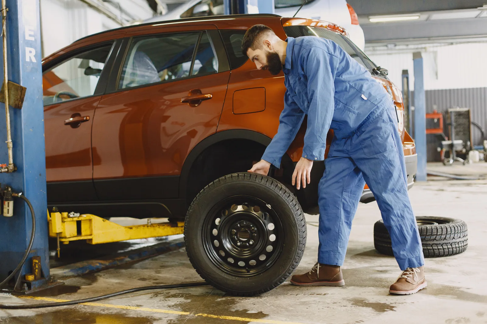 A mechanic in blue coveralls fits a wheel to a car raised on a workshop lift.