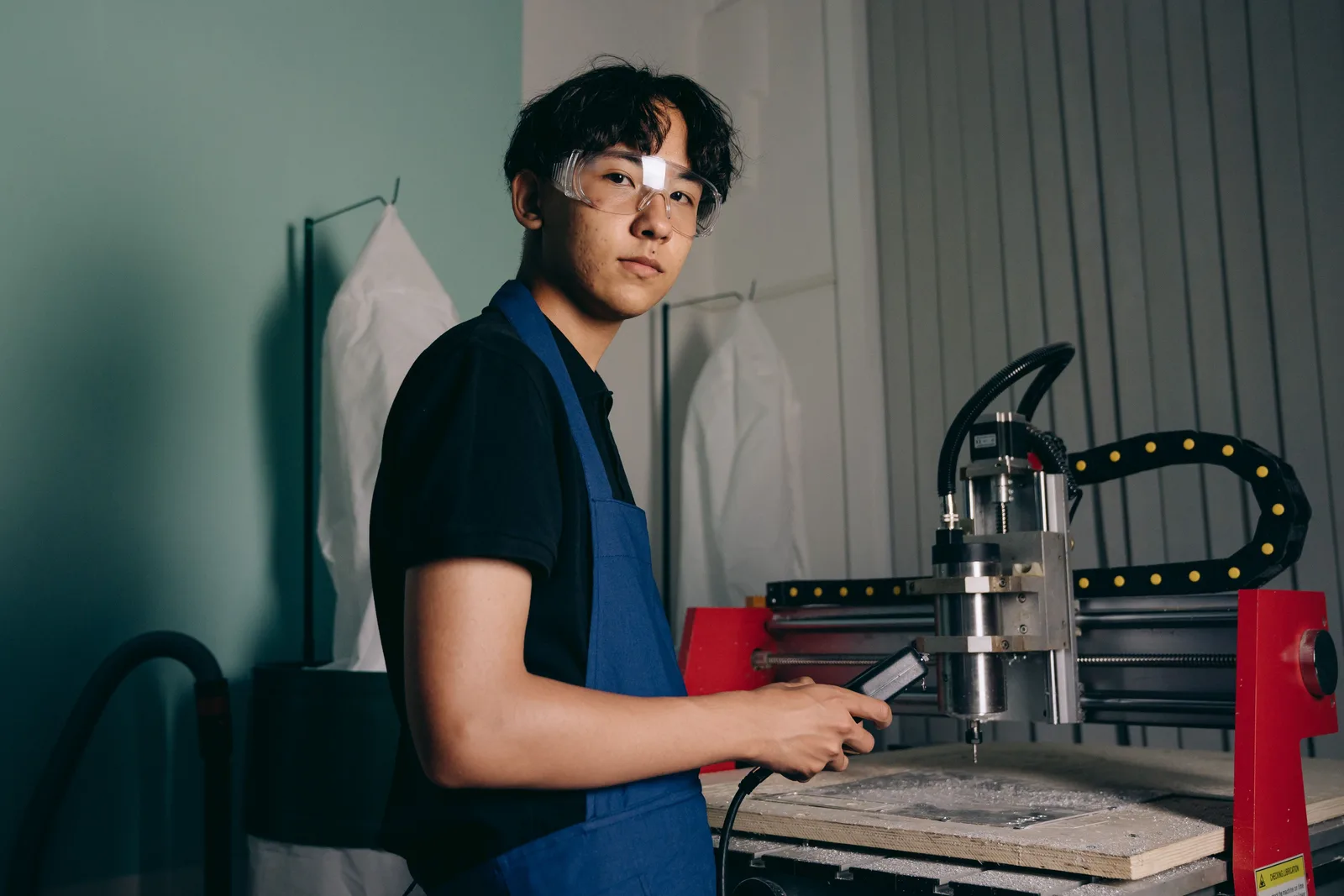 A machinist in safety glasses operates a CNC router at a bench in a small workshop