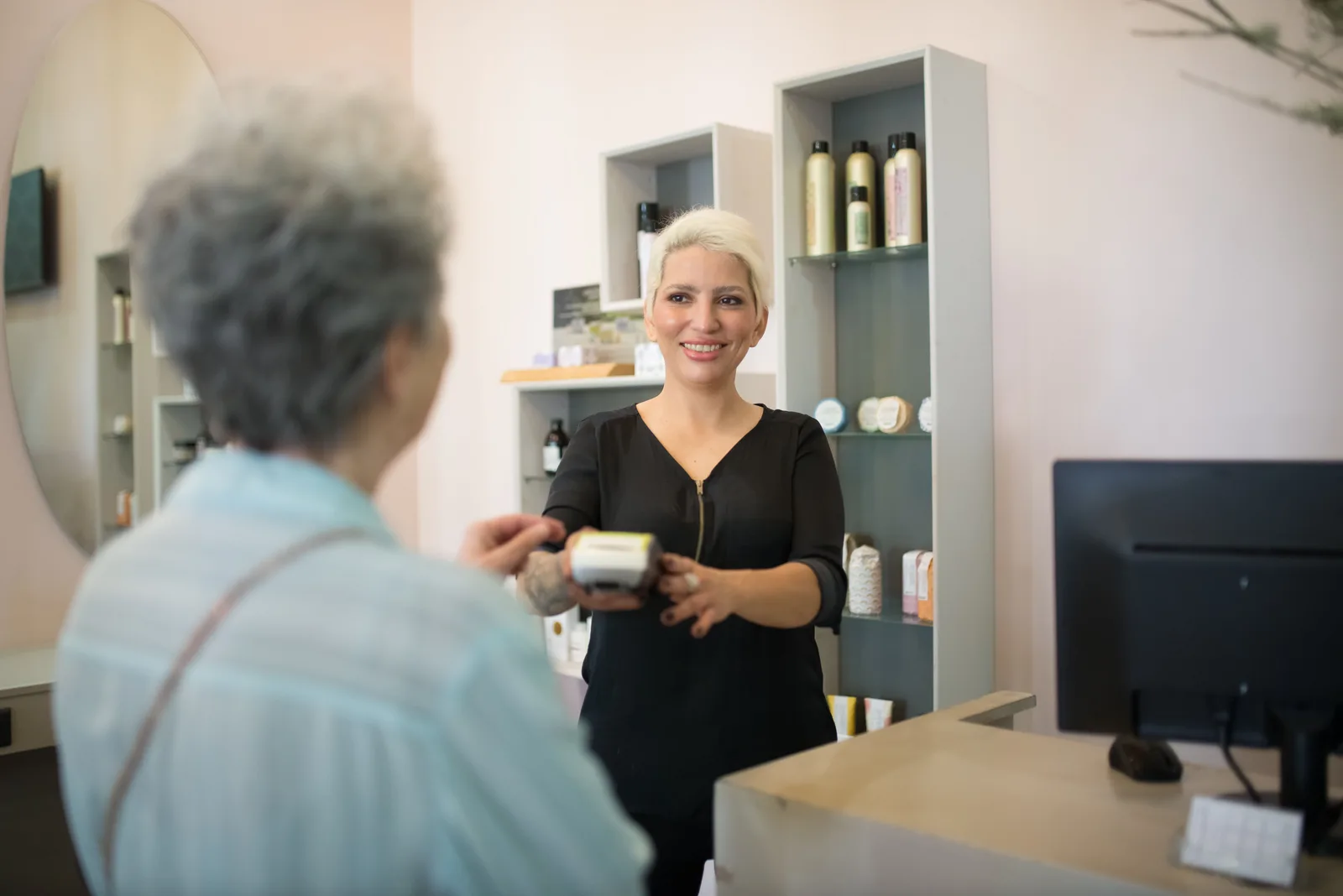 A salon owner in black handing a card reader to a client at the reception desk, with product shelves behind