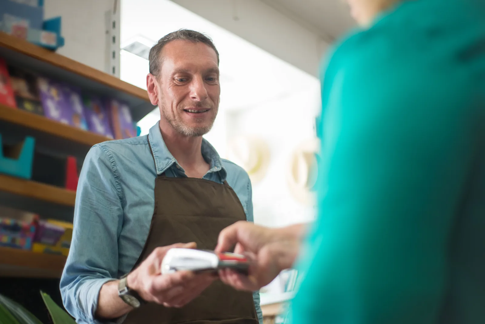 A shopkeeper in an apron holding out a card terminal to a customer across the counter of a small store