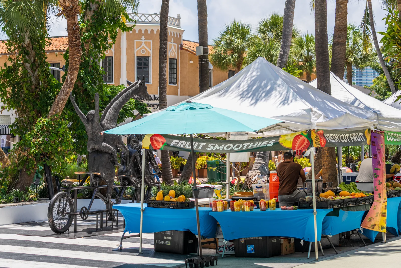 Open air market stalls under white canopies selling fresh cut fruit and smoothies, with palms and a tiled roof building behind