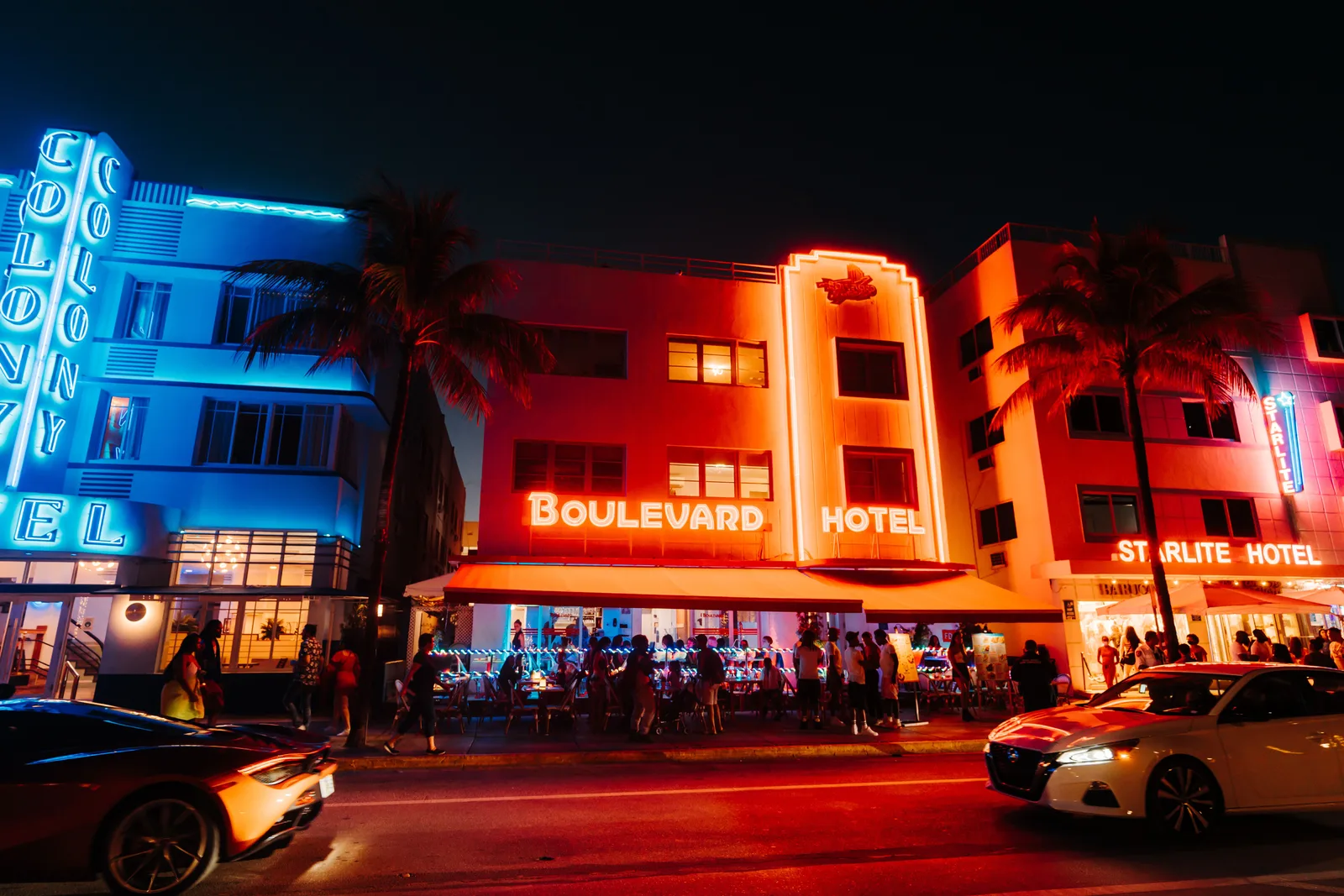 Neon lit Art Deco hotels along Ocean Drive at night with full sidewalk restaurant terraces and traffic passing