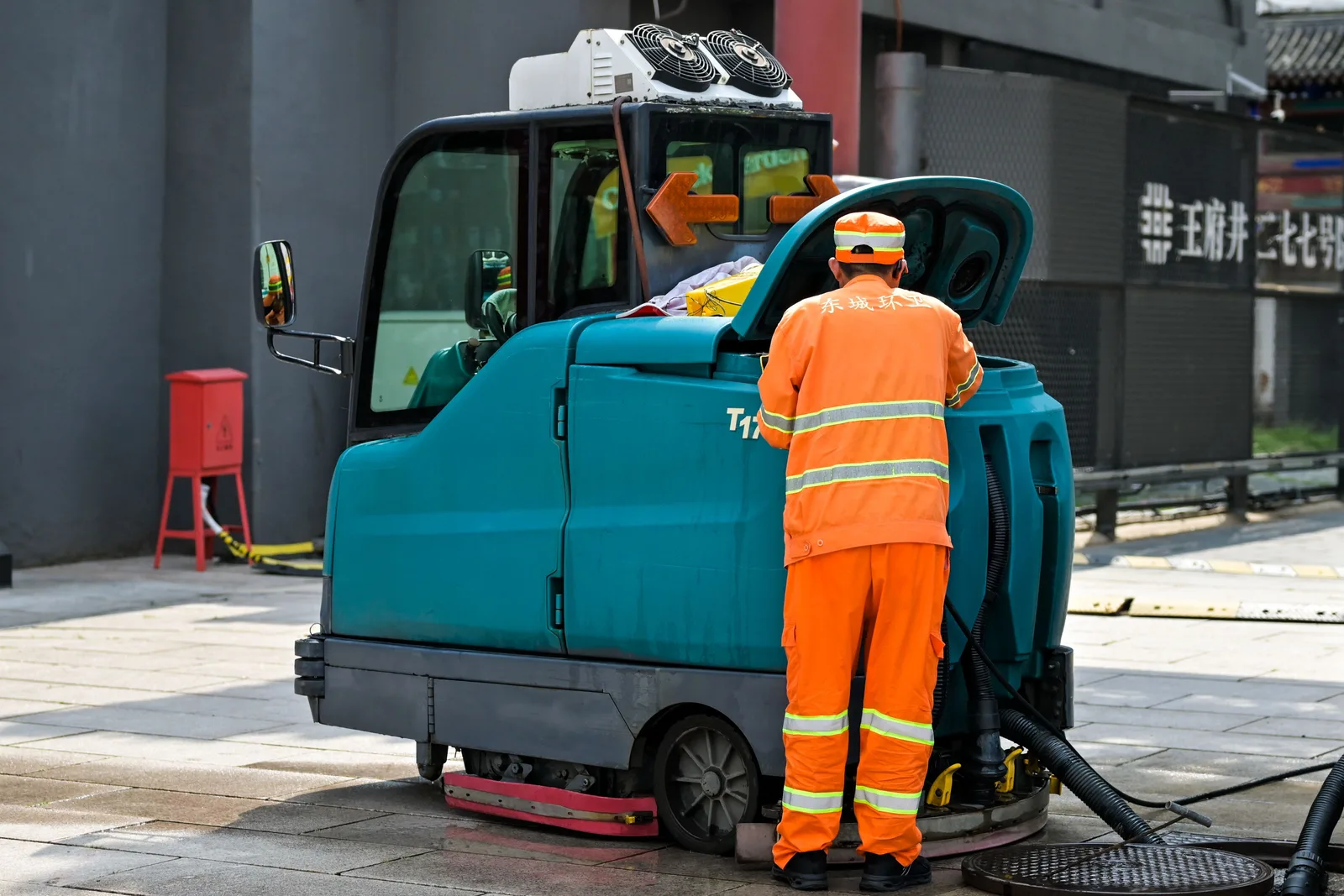 A uniformed street cleaner walking behind a powered sweeping machine on a city pavement.