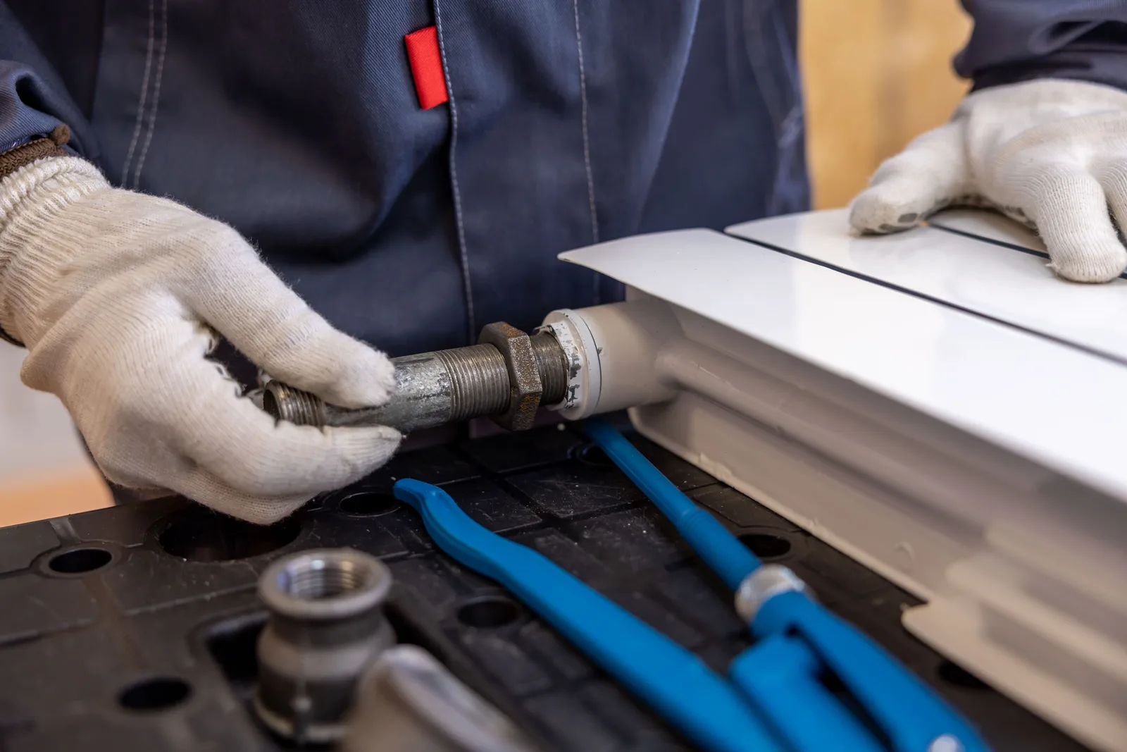 A plumber in gloves fitting a valve onto a white radiator, with a pipe wrench on the bench alongside