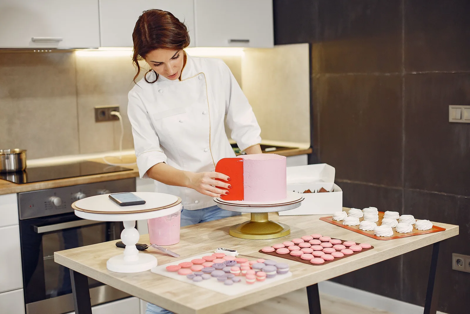 A pastry chef in whites finishing a decorated cake at a work table set out with trays of macarons.