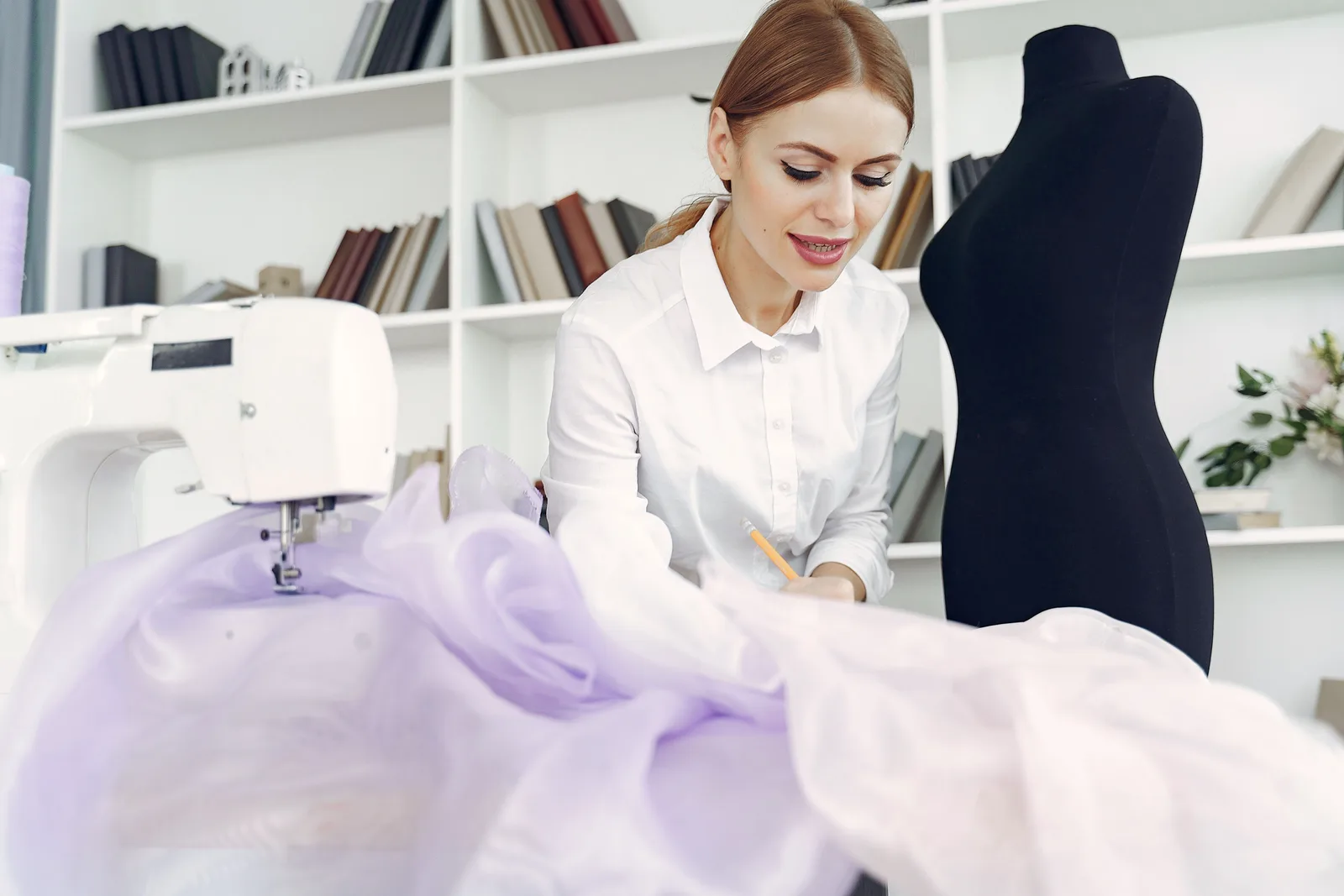 A dressmaker working fabric at a table beside a sewing machine and a dress form in her studio.