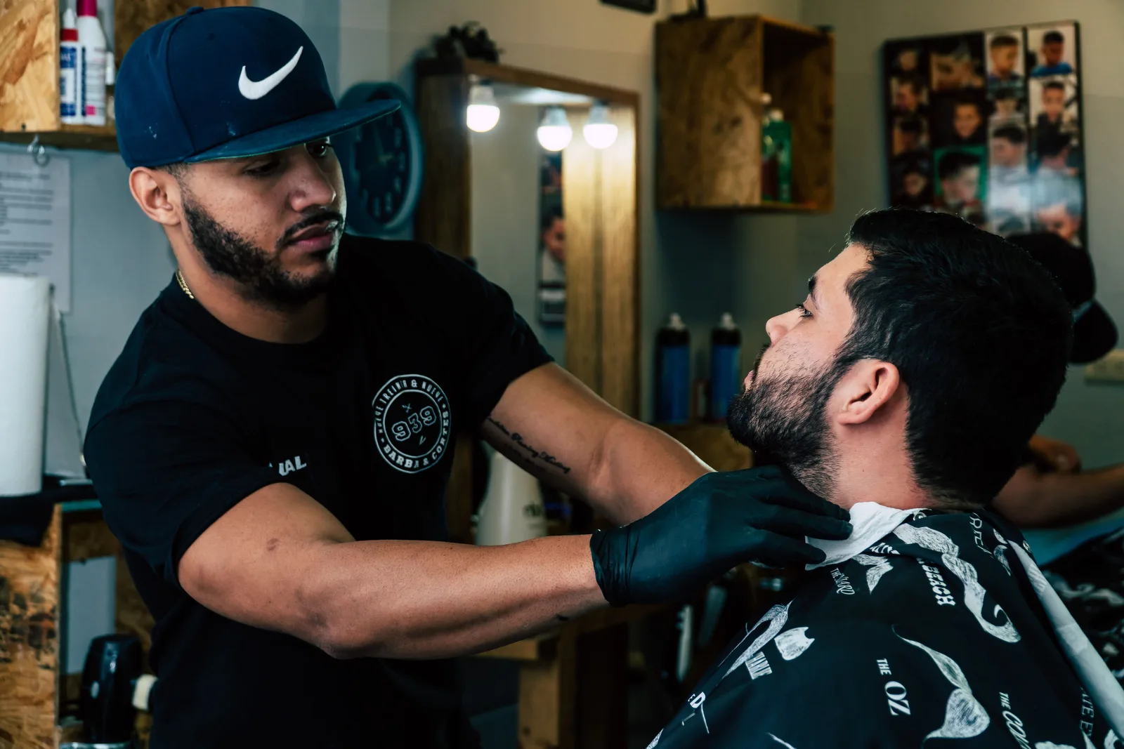 A barber in a shop tee shirt trimming a seated client's beard, with mirrors and product shelves behind