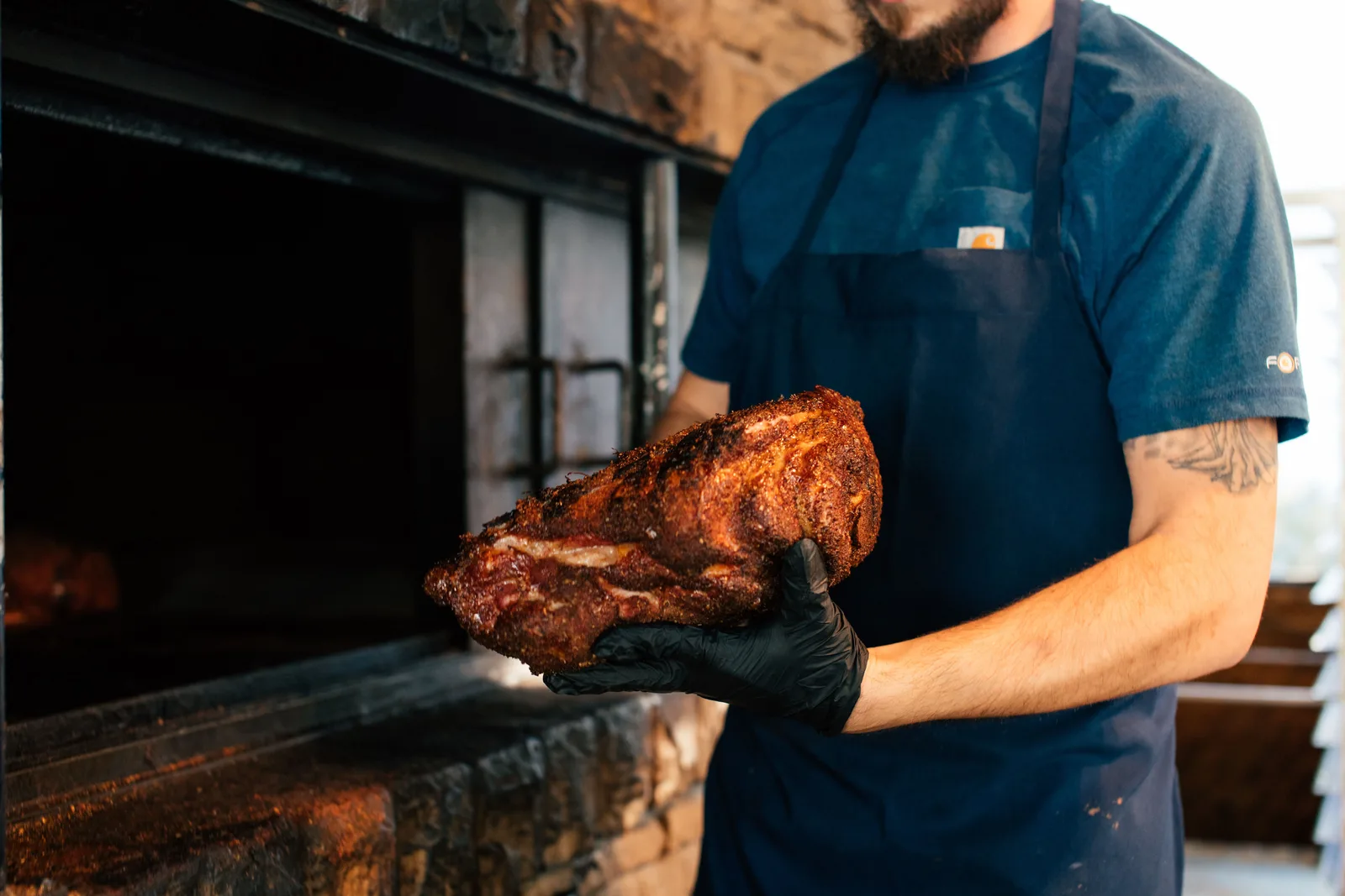 A cook in an apron and black gloves holding a smoked pork shoulder beside a brick barbecue pit