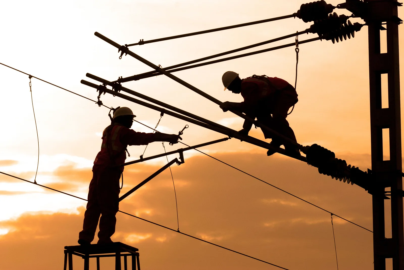 Two electricians silhouetted against a sunset while working on overhead line equipment