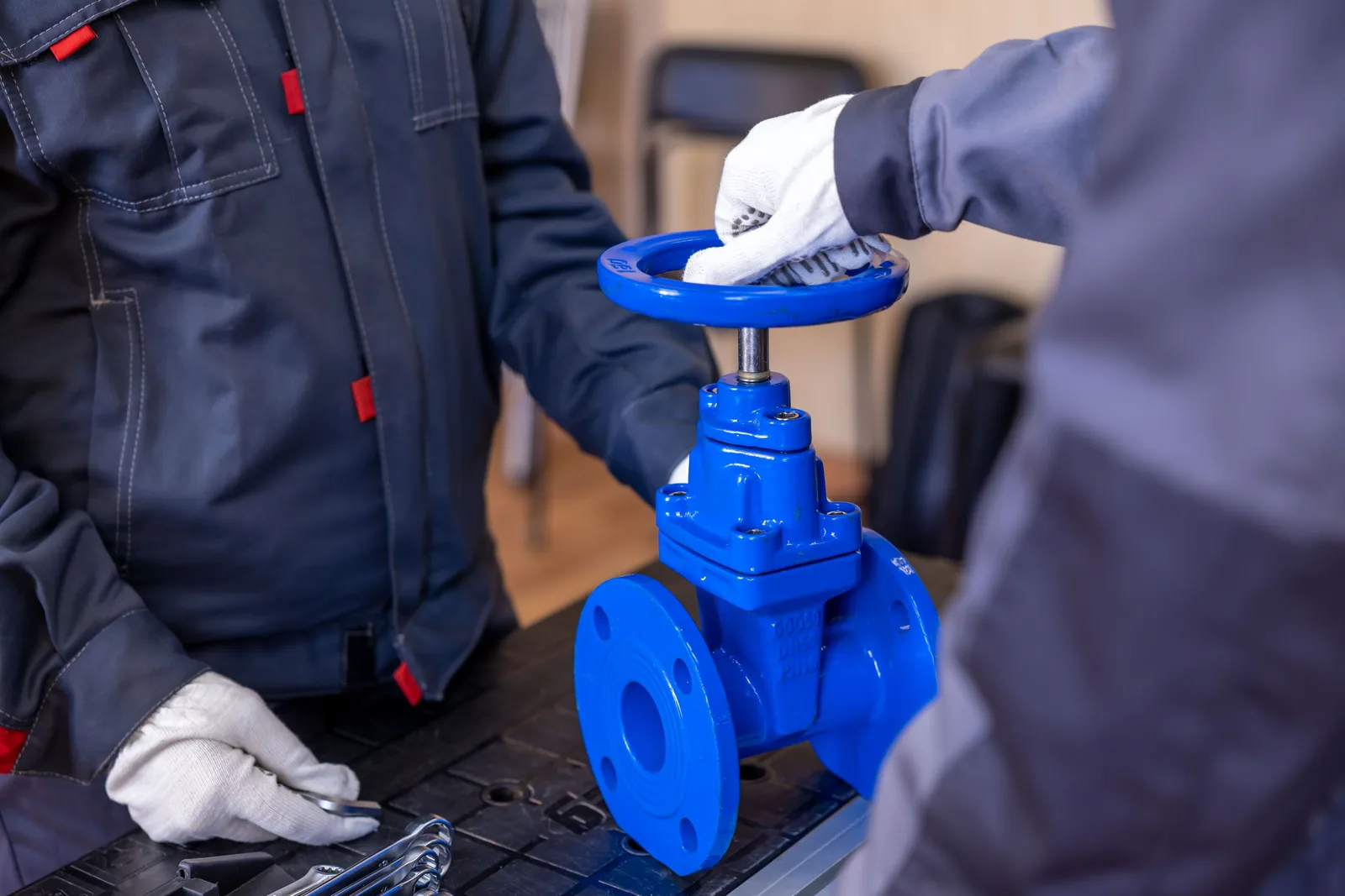 Two workers in overalls handling a large blue flanged gate valve on a bench, with wrenches laid out beside them