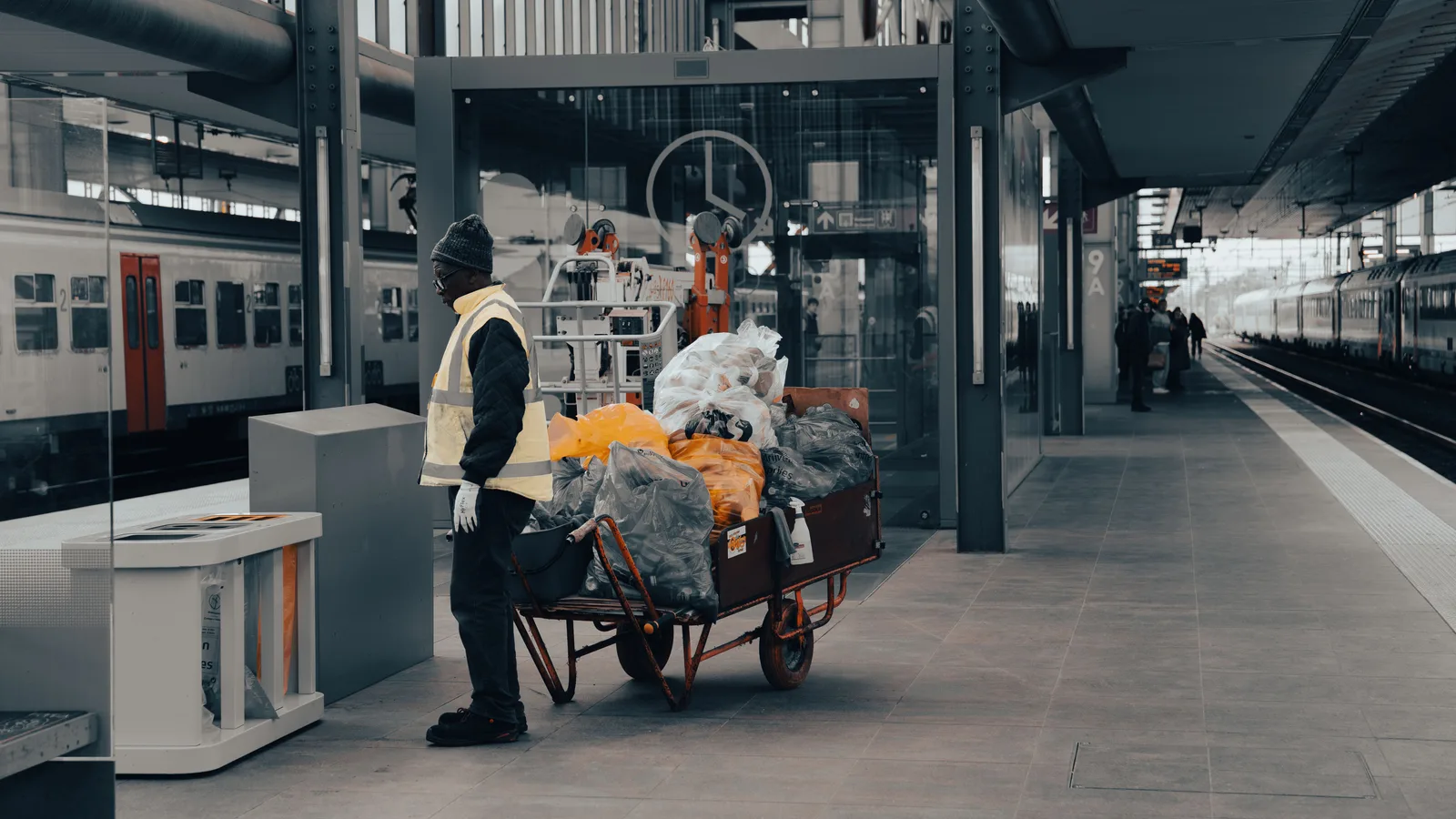 A worker in a high visibility vest pushing a cart loaded with bagged waste along a station platform.