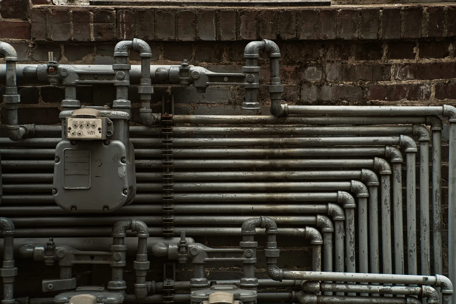 A dense bank of meters and pipework mounted on the brick wall of an older multi-unit building