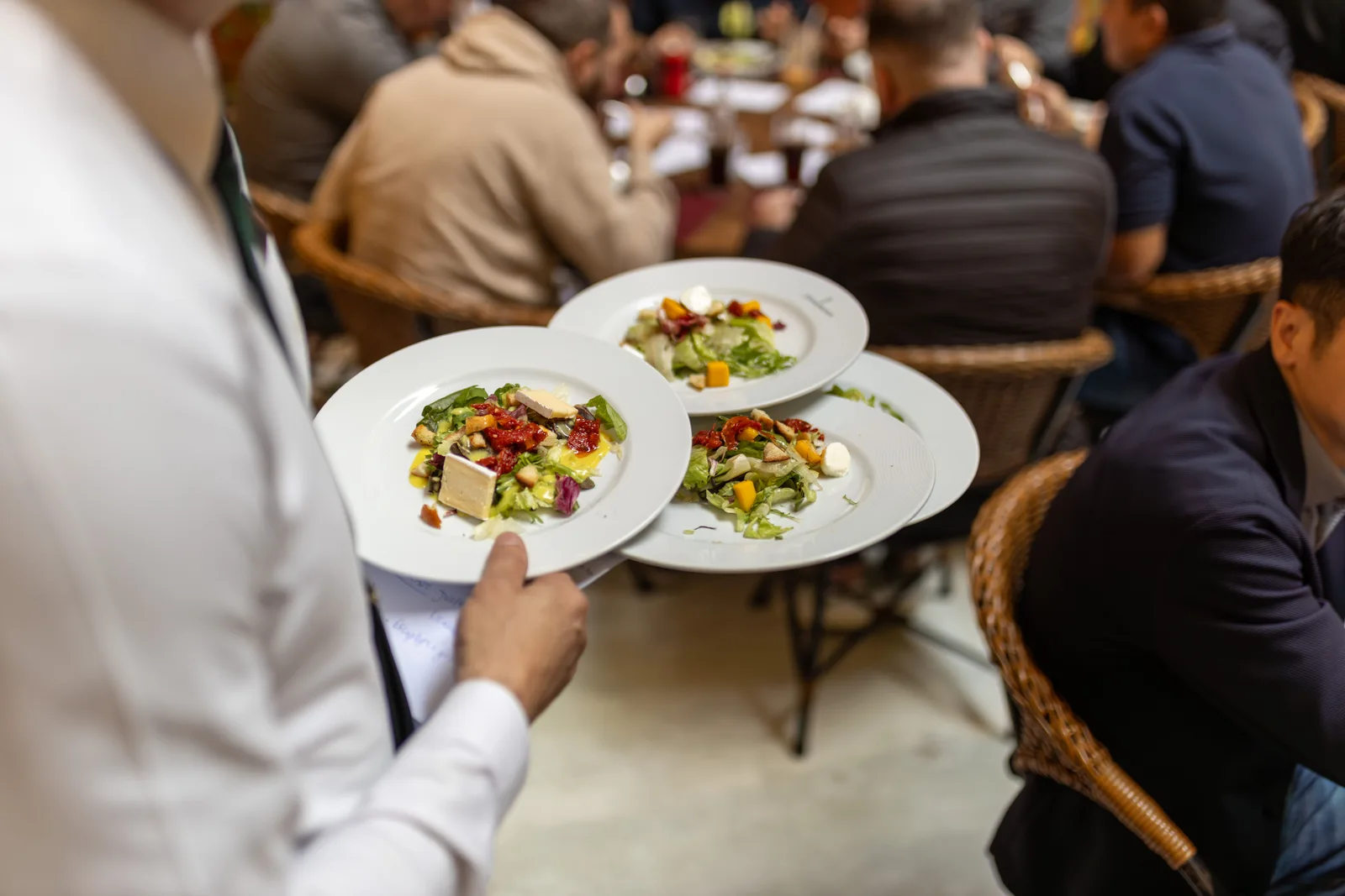 Server carrying three plated salads through a crowded dining room