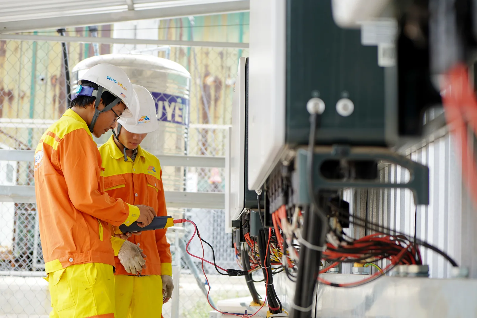 Dos técnicos con ropa de alta visibilidad trabajando en el cableado dentro de una unidad eléctrica al aire libre