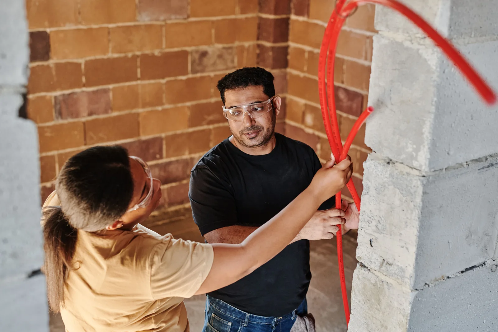 Two workers feeding red flexible tubing through an opening in a block wall inside a building under construction
