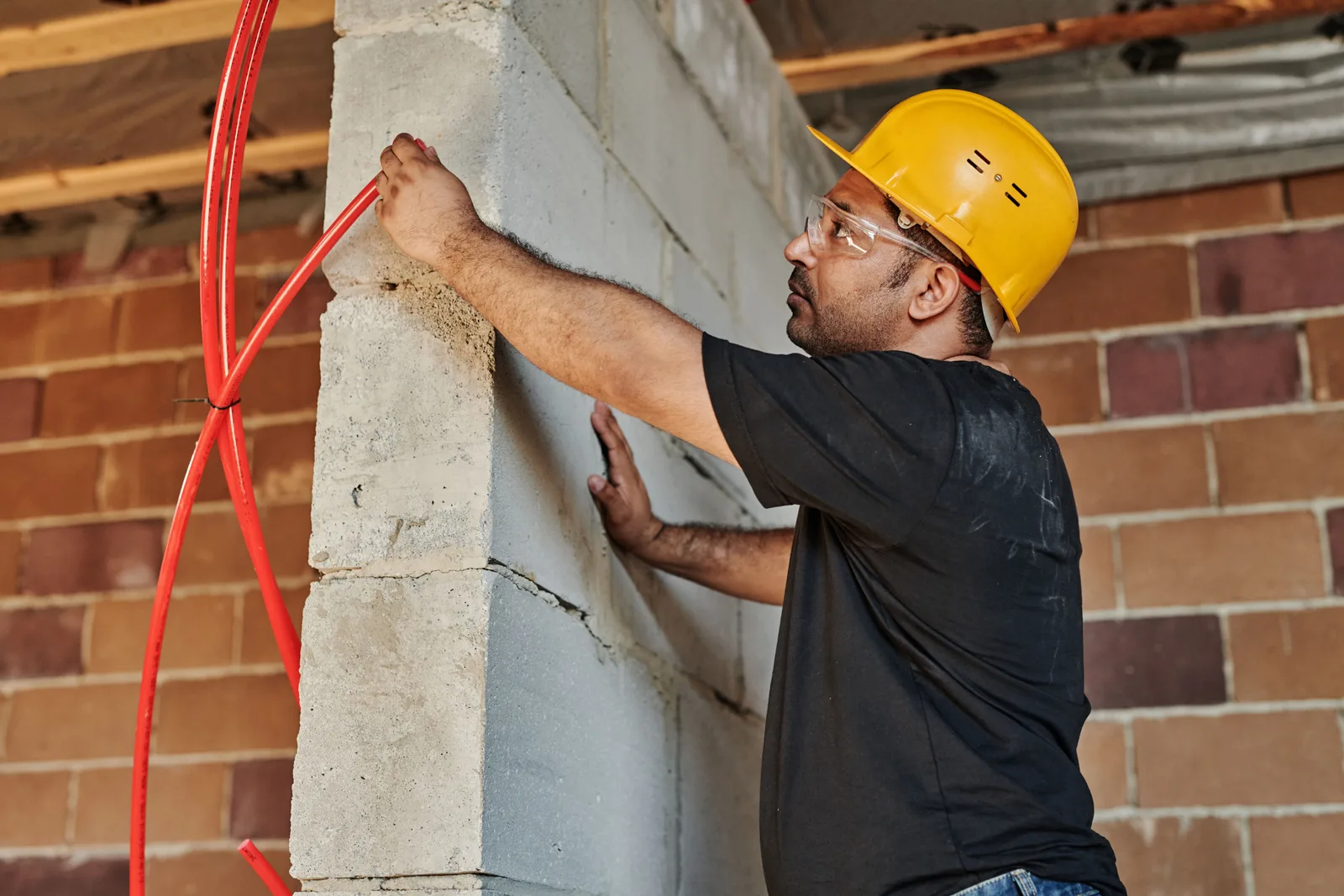 An electrician in a yellow hard hat fixing red conduit to a wall on a construction site