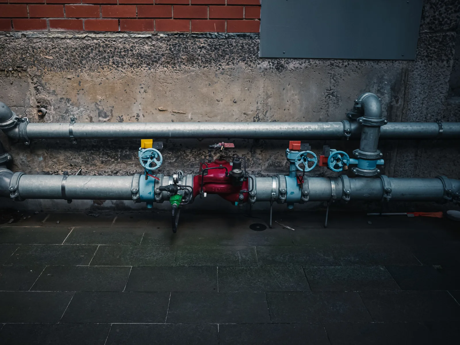 A run of pipework with colored valve handles mounted along the base of a building wall